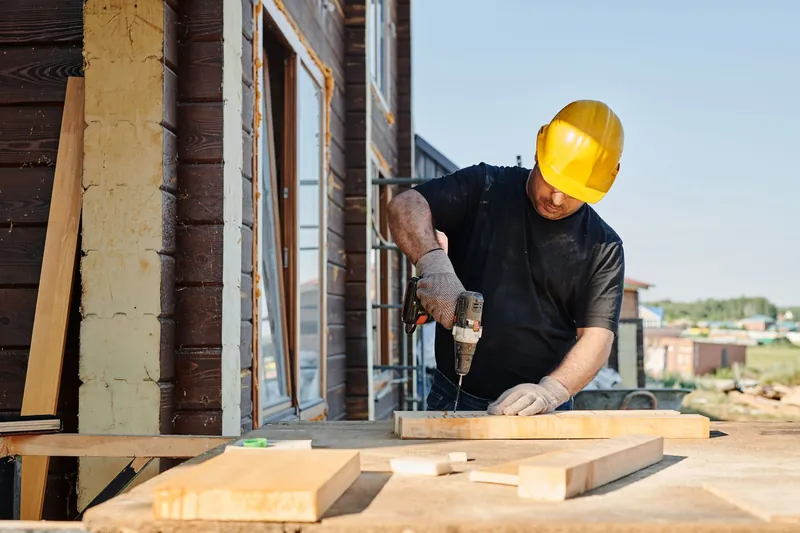 Contractor in a hard hat and work gloves drilling lumber on a job site