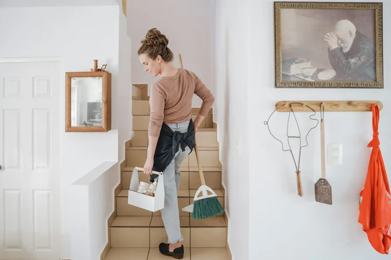 Cleaner carrying a supply caddy and broom up the stairs of a home